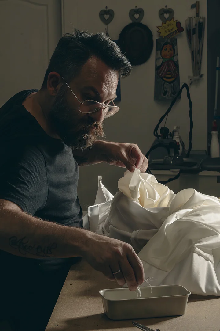 Hombre con barba cosiendo tela blanca en un taller de costura. - Gianluca Urraso